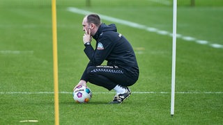 Ole Werner hockt auf dem Boden, hält dabei eine Hand auf einen Ball und schaut nachdenklich.