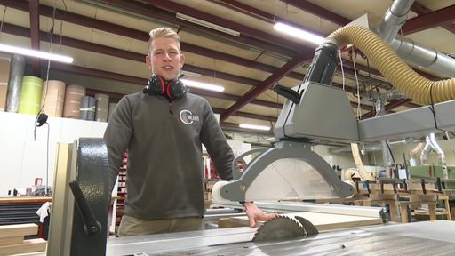 Jonas Kamermann in einer Tischler-Werkstatt. Vor ihm steht eine Maschine mit der scheinbar Holz bearbeitet wird.