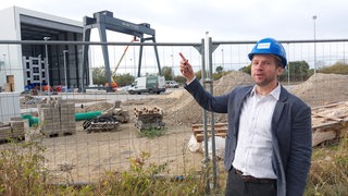 Ein Mann mit Helm zeigt auf eine Halle.